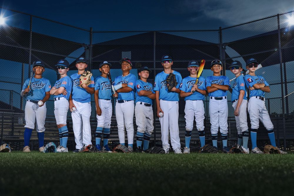 Youth baseball team poses together in light blue uniforms on a field at dusk for team photo.