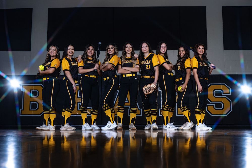 Basketball team wearing black and yellow uniforms poses in front of illuminated BULLS lettering on dark stage.