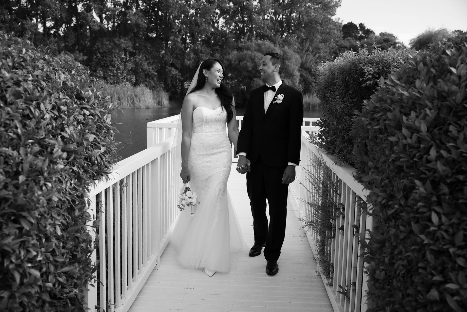 A black and white wedding photo of a couple walking together on a white railing bridge surrounded by lush greenery.