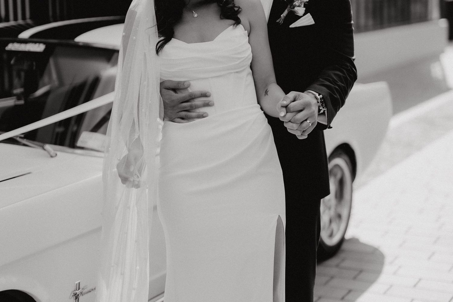 A close-up black and white photo of a couple embracing next to a vintage car.
