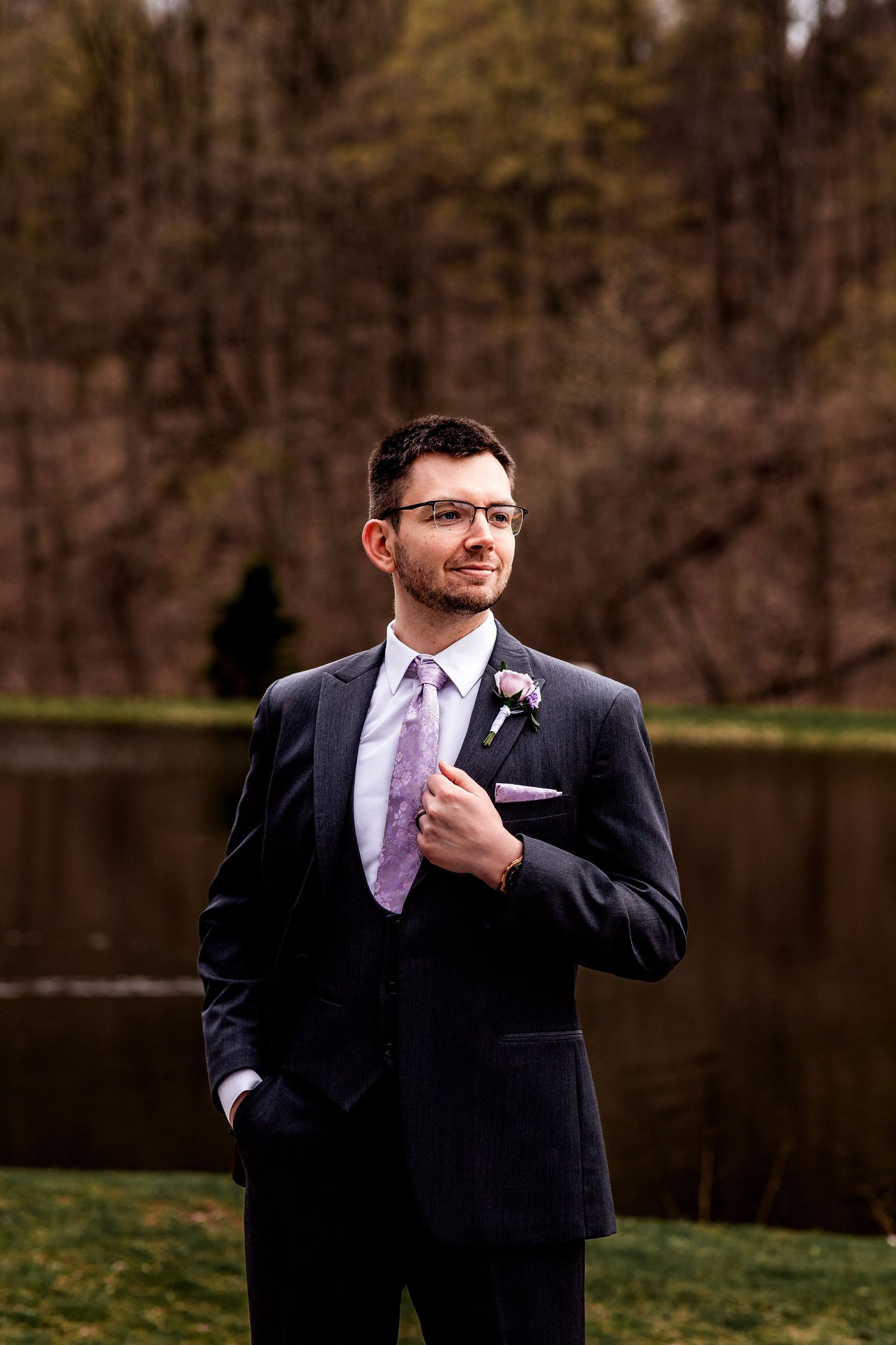 A person in a dark formal suit with glasses and purple tie poses outdoors during what appears to be a wedding event.