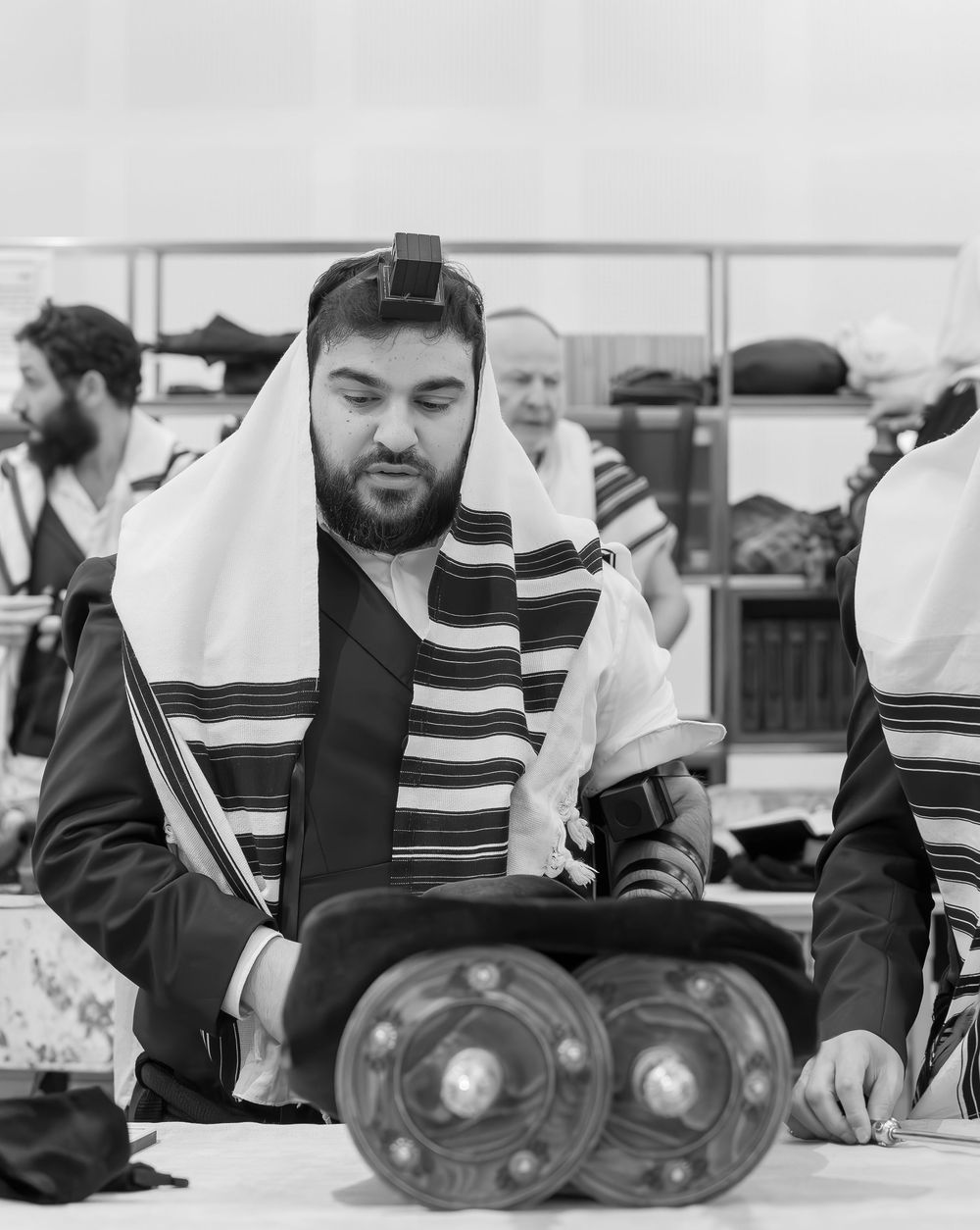 A person in traditional Jewish prayer attire reads from prayer texts during a religious service in black and white.