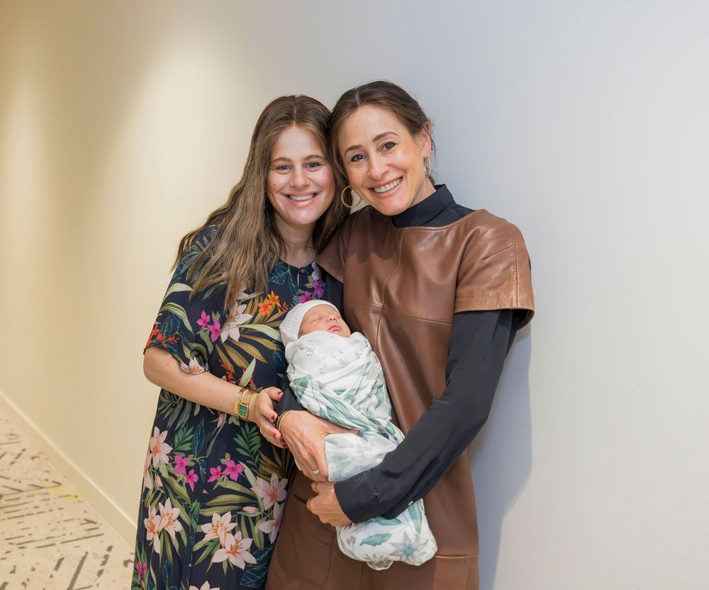 Two people holding a swaddled baby stand together against a beige wall, one wearing a floral dress.