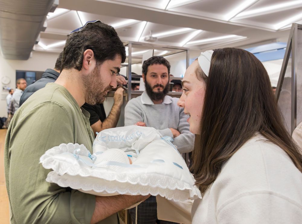 Family gathers around a newborn baby swaddled in white blanket inside a bright hospital room.
