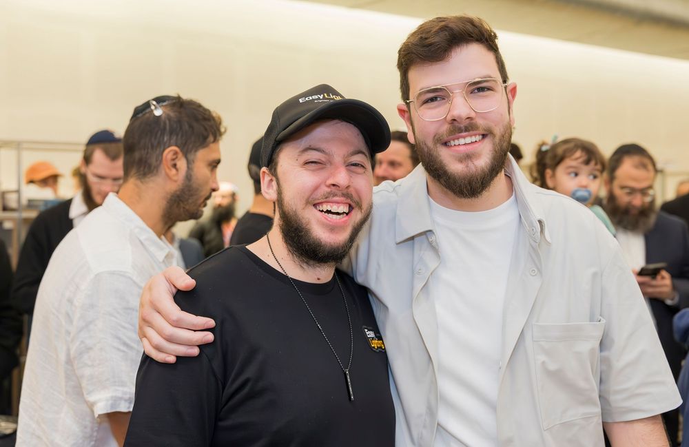Two people sharing a friendly moment together at an indoor event with other guests in the background.