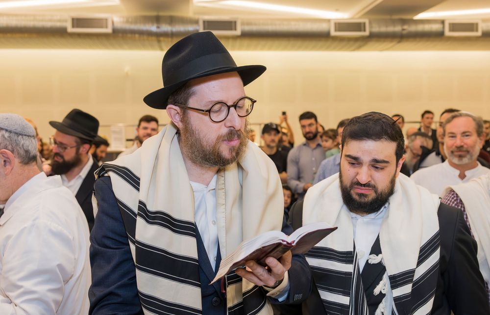 Group of Orthodox Jewish men wearing prayer shawls and hats gathered in prayer at a religious service.