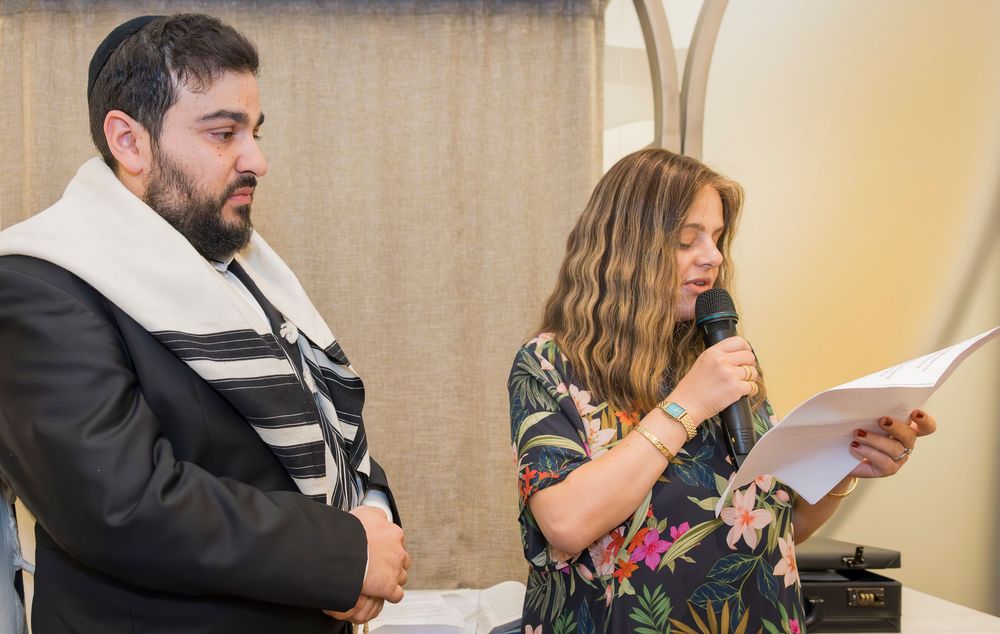 A person in a patterned dress speaks into a microphone while reading from papers next to someone in religious attire.