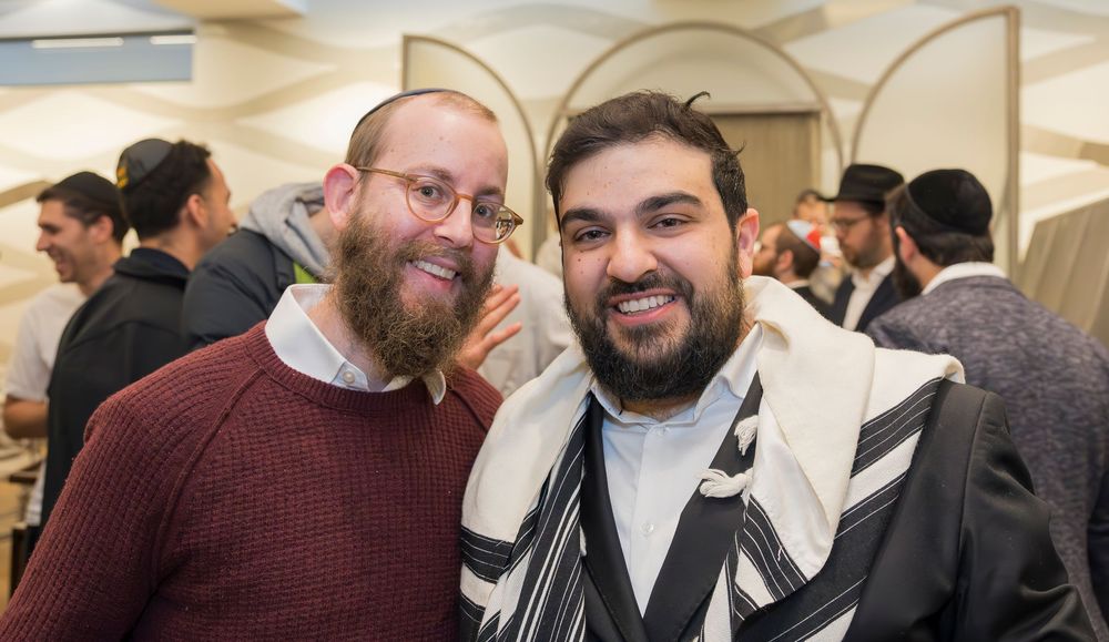 Two people wearing religious garments smile together at a social gathering with others visible in the background.