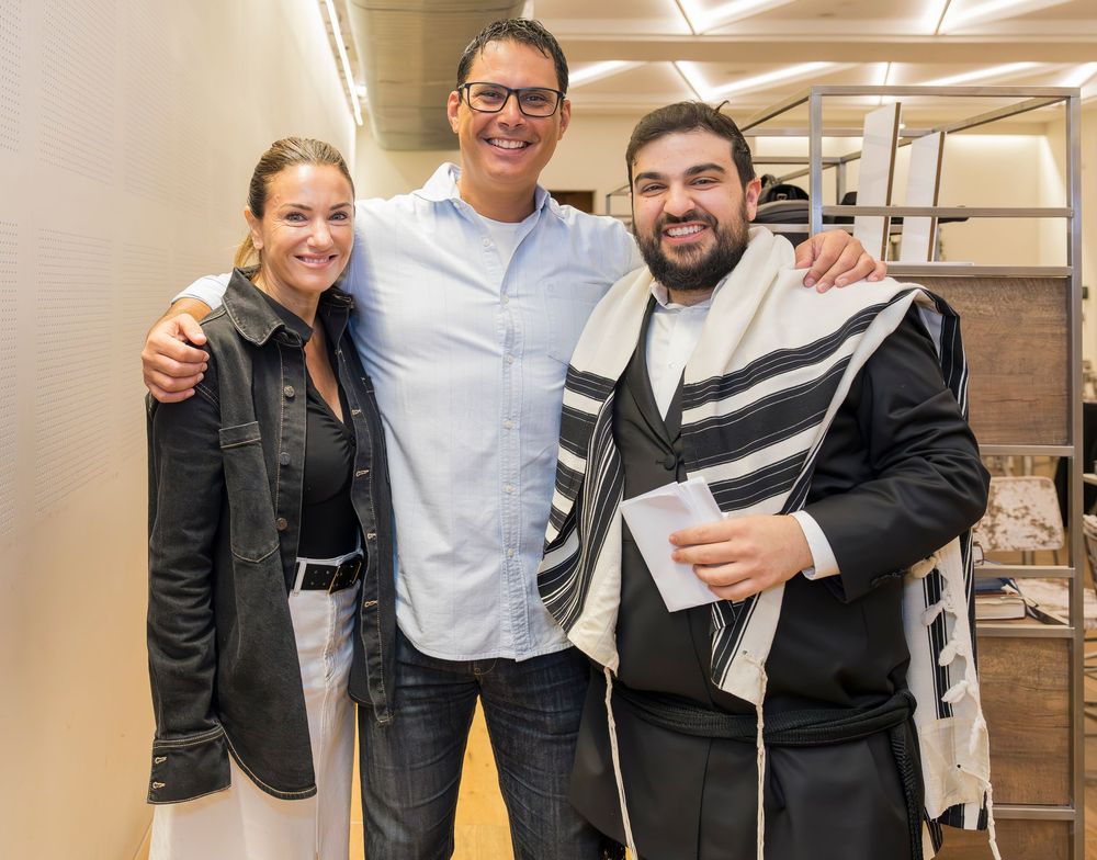 Three people stand together smiling in an office hallway, wearing casual attire and winter jackets.