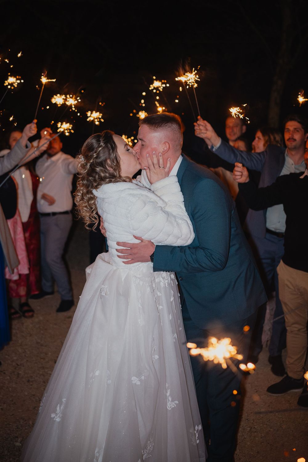 Newlyweds share an intimate kiss at nighttime wedding celebration surrounded by guests holding sparklers.