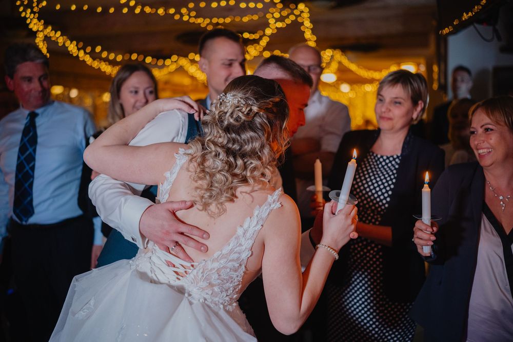 A couple embraces on a dimly lit dance floor with string lights overhead while guests hold candles during a wedding reception.