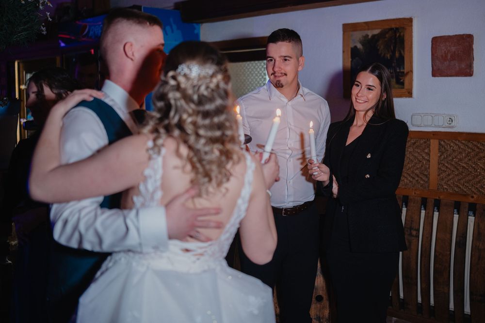 A couple in wedding attire chats with guests during an evening reception at a rustic venue.