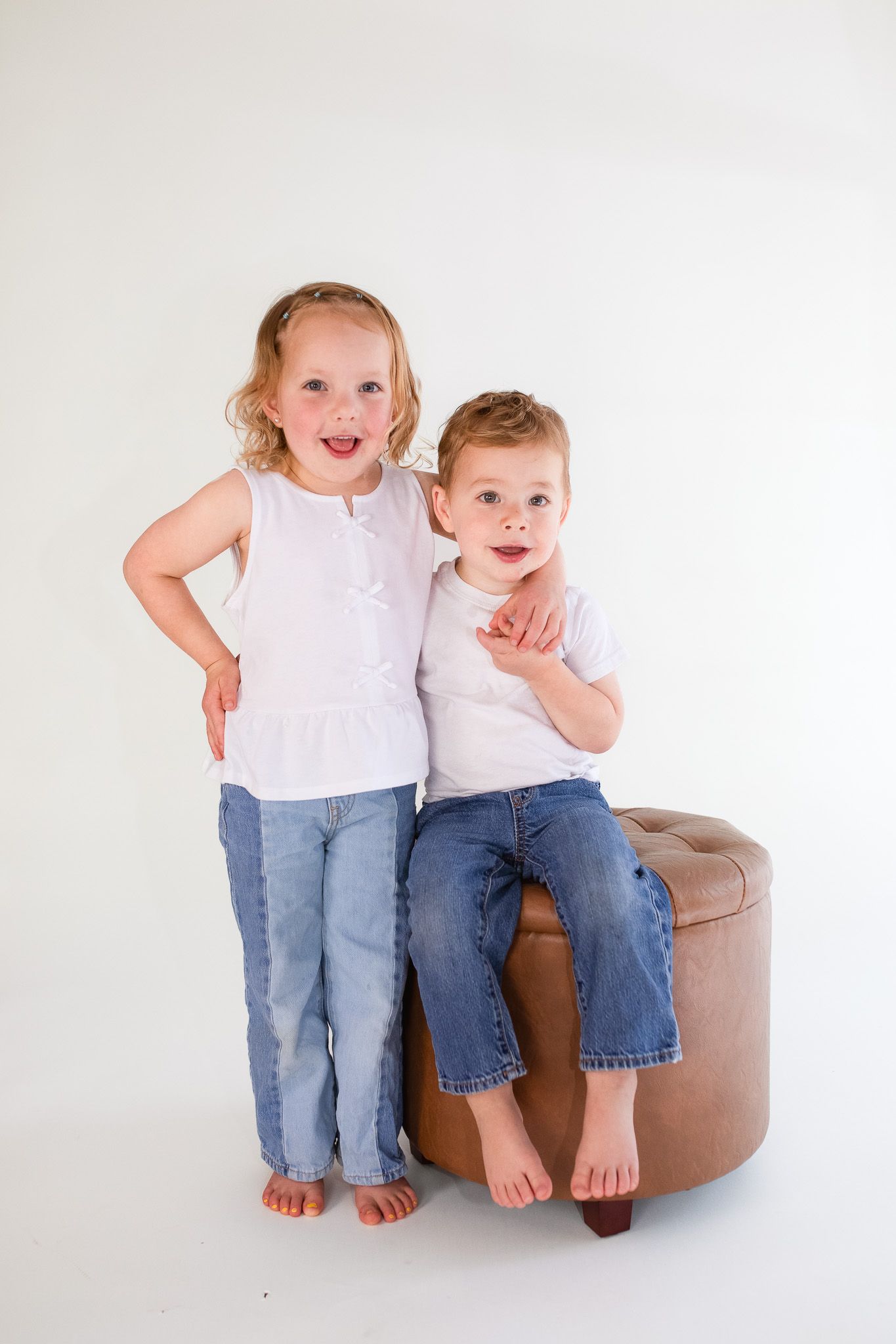 Two young children in blue jeans and white tops pose together while sitting on a brown leather ottoman against white backdrop.