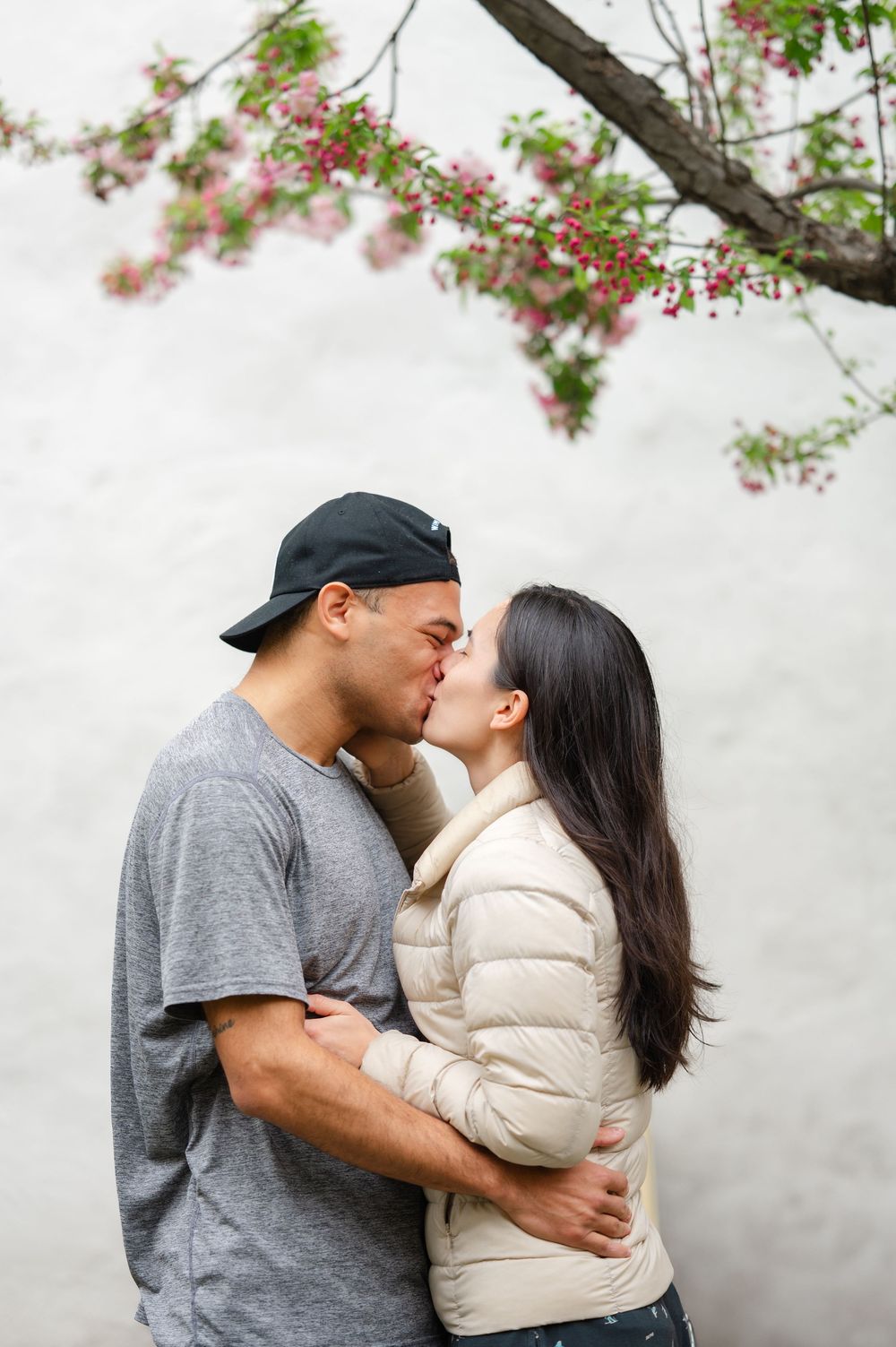 Couple kissing beneath blooming cherry blossoms against a white wall outdoors during an engagement session in NYC