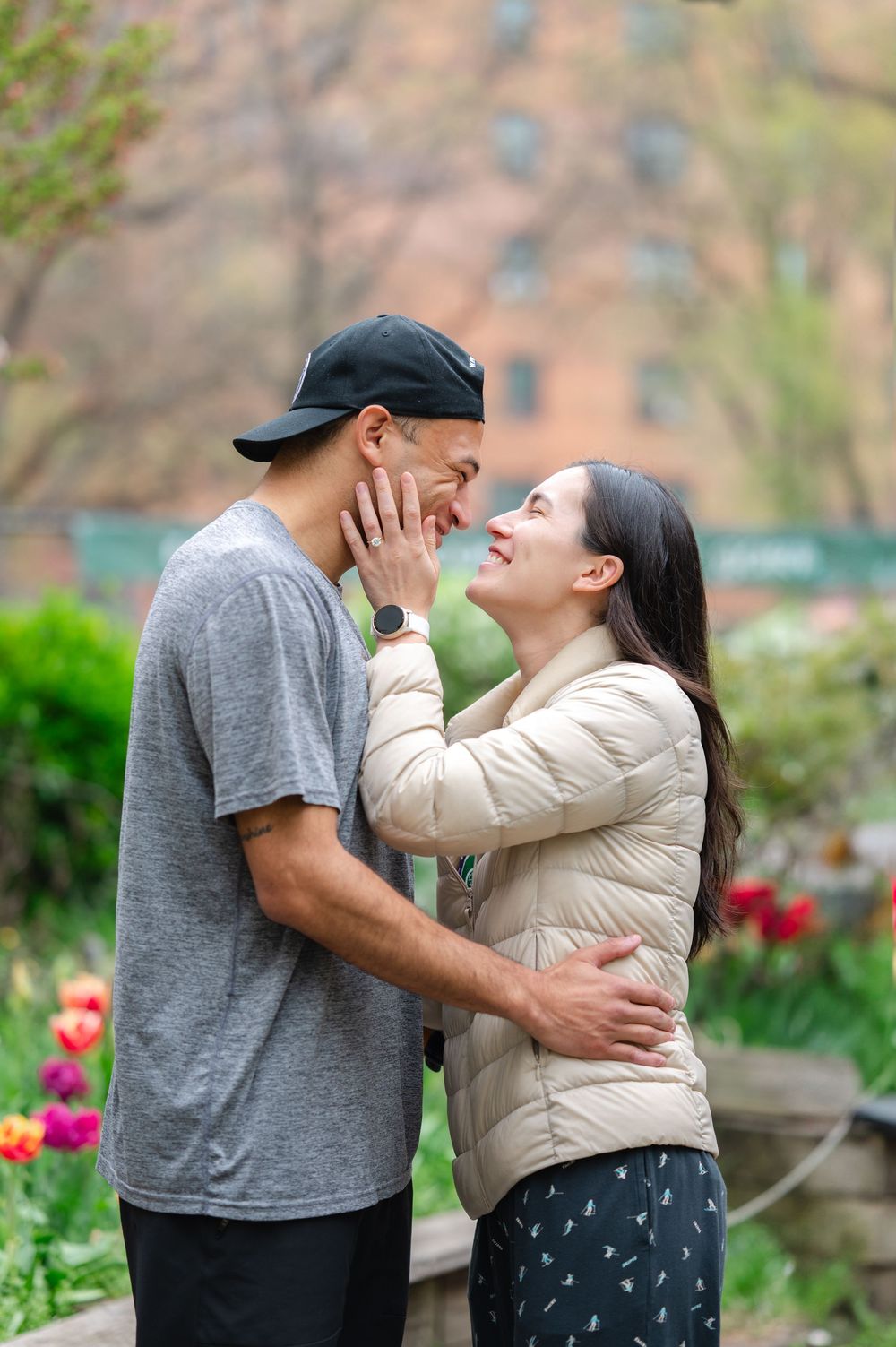 A loving couple embrace in a garden with blooming pink tulips in the background after their proposal in on the Upper West Side of NYC