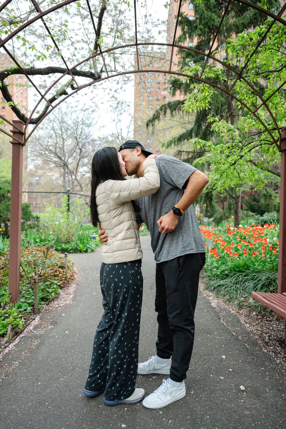 A couple shares a romantic kiss under an arched trellis path surrounded by spring flowers and greenery after their proposal moment.