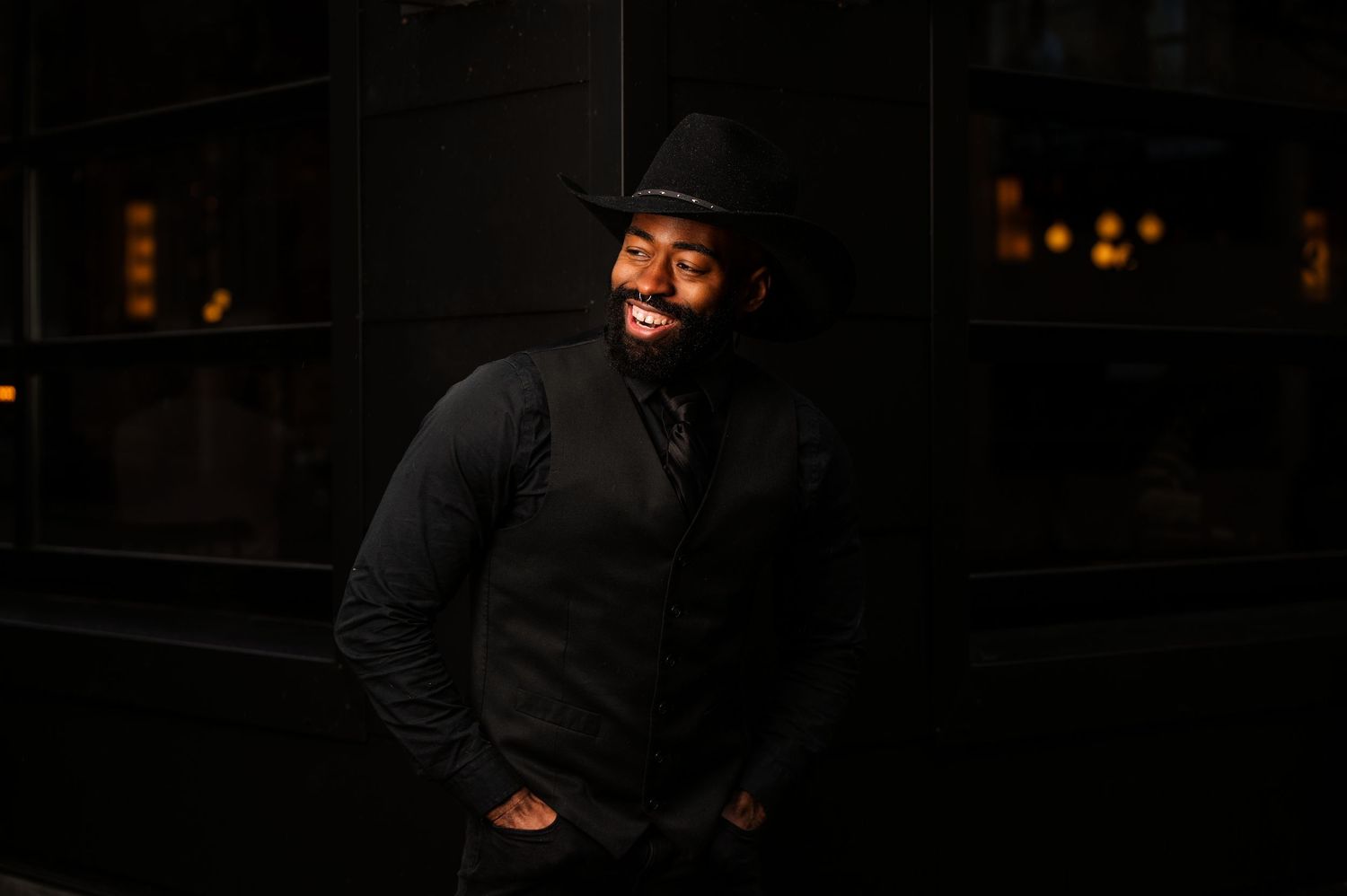 Man in black outfit and cowboy hat smiling confidently in moody lighting. Photo by Carolina Waldroff.