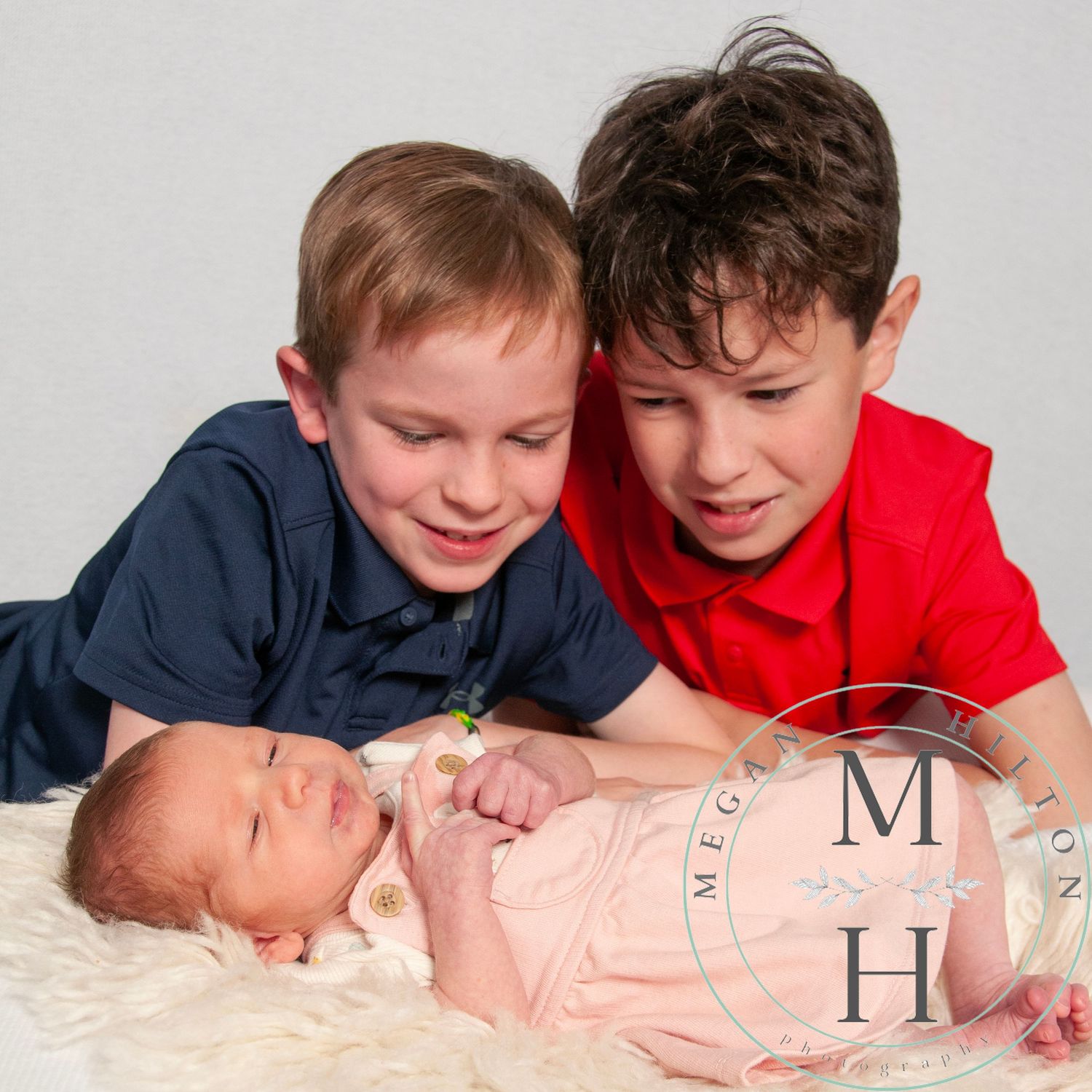 Two boy siblings looking adoringly at their sleeping newborn sister on white fur bedding.