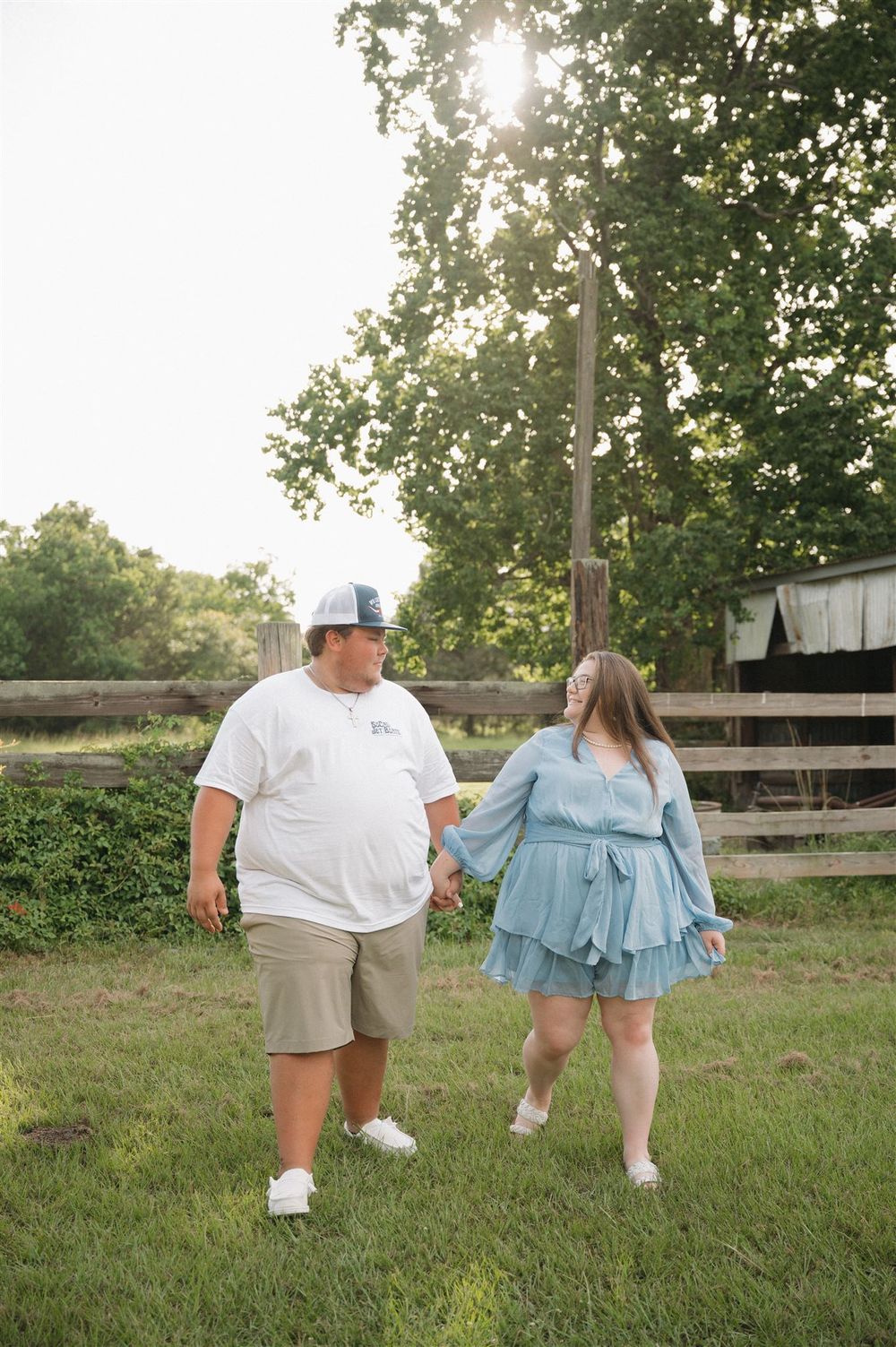 Two people holding hands while walking in a grassy field with wooden fences and trees in the background at sunset.