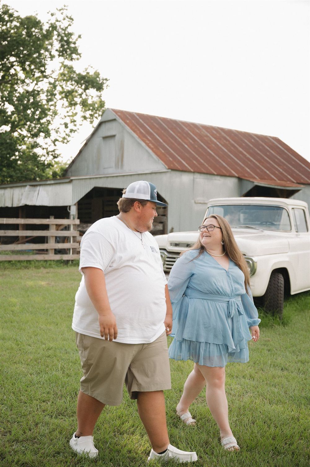 A couple shares a joyful moment together in front of a rustic barn and vintage truck on a grassy lawn.