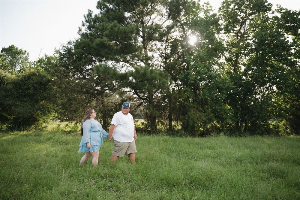 Two people in light colored clothing stand together in a sunlit grassy field with trees in the background.