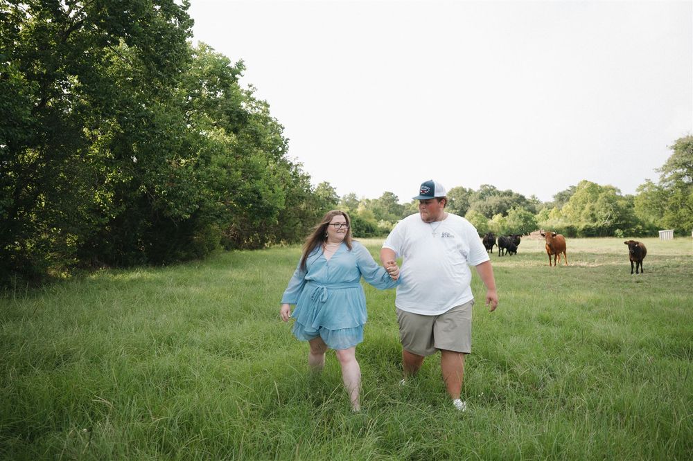 Two people walk hand in hand through a sunny green meadow with grazing horses visible in the background.
