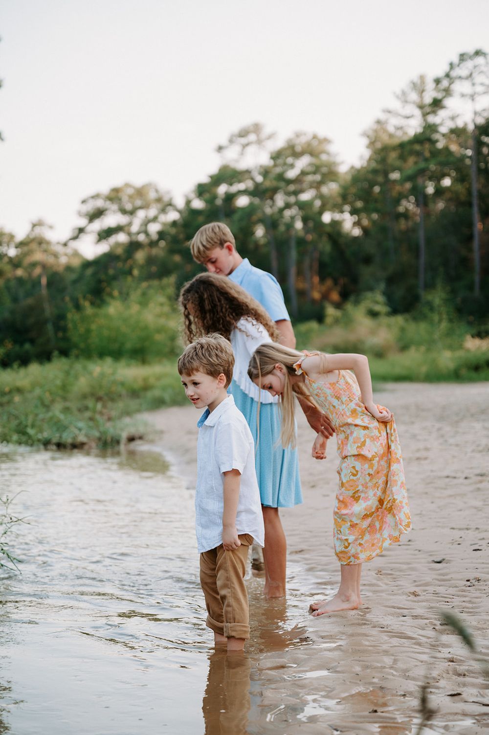 Three young people explore together at the water's edge on a summer afternoon with trees in the background.