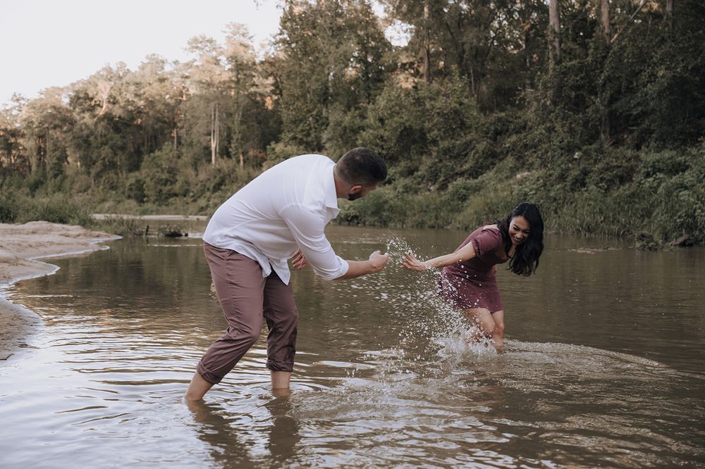 Two people splash and play together in a shallow river surrounded by lush forest scenery during daytime.