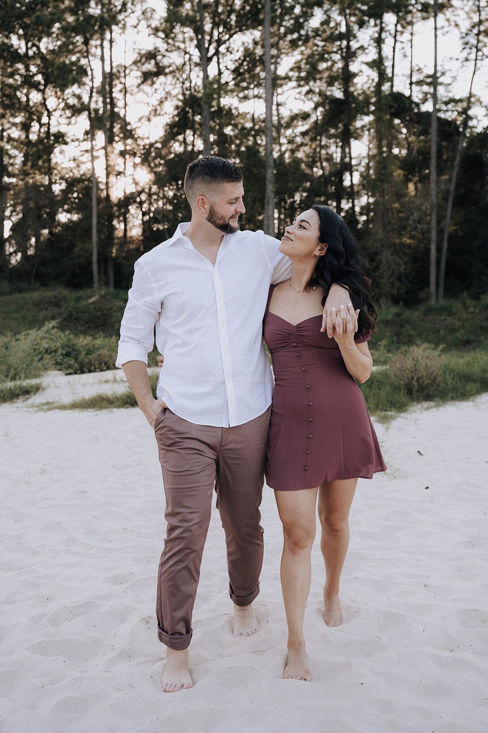 A couple walks barefoot on a sandy beach path together at sunset with tall pine trees in the background.