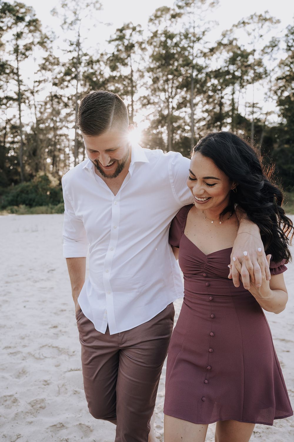 A romantic couple walking together on a sandy beach at sunset, with tall pine trees in the background.