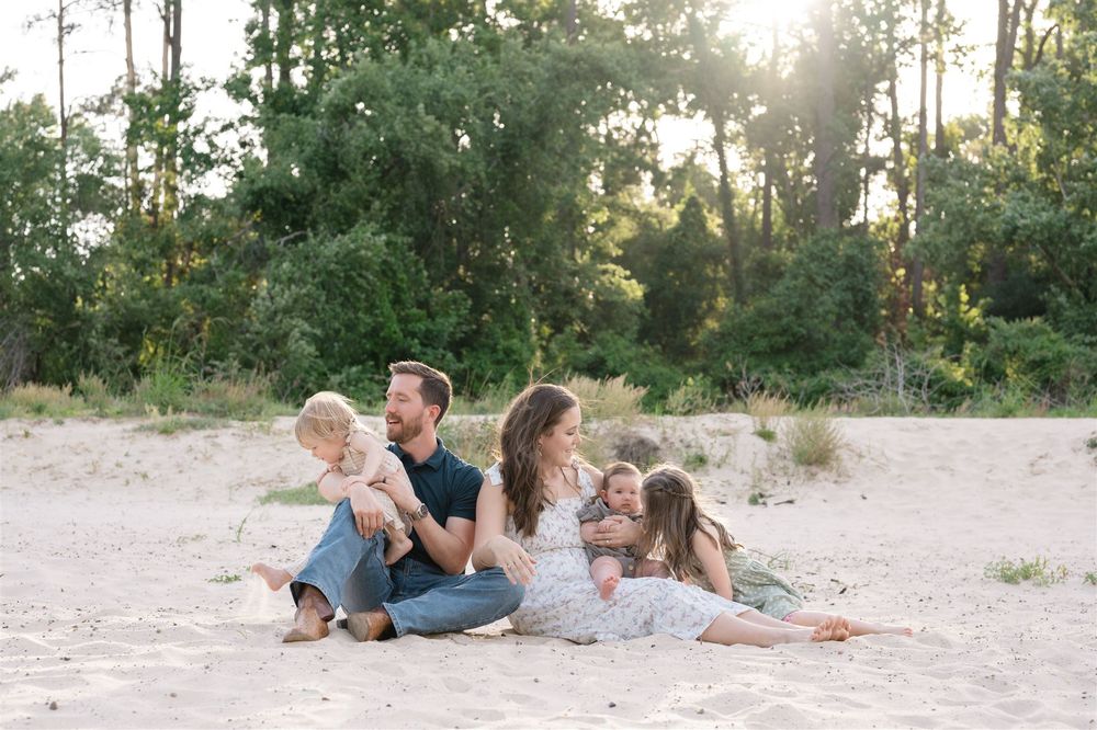 A family sits together on a sandy beach at sunset with pine trees in the background.