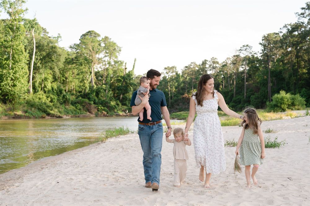 Family walks along sandy riverbank with trees in background on summer day.