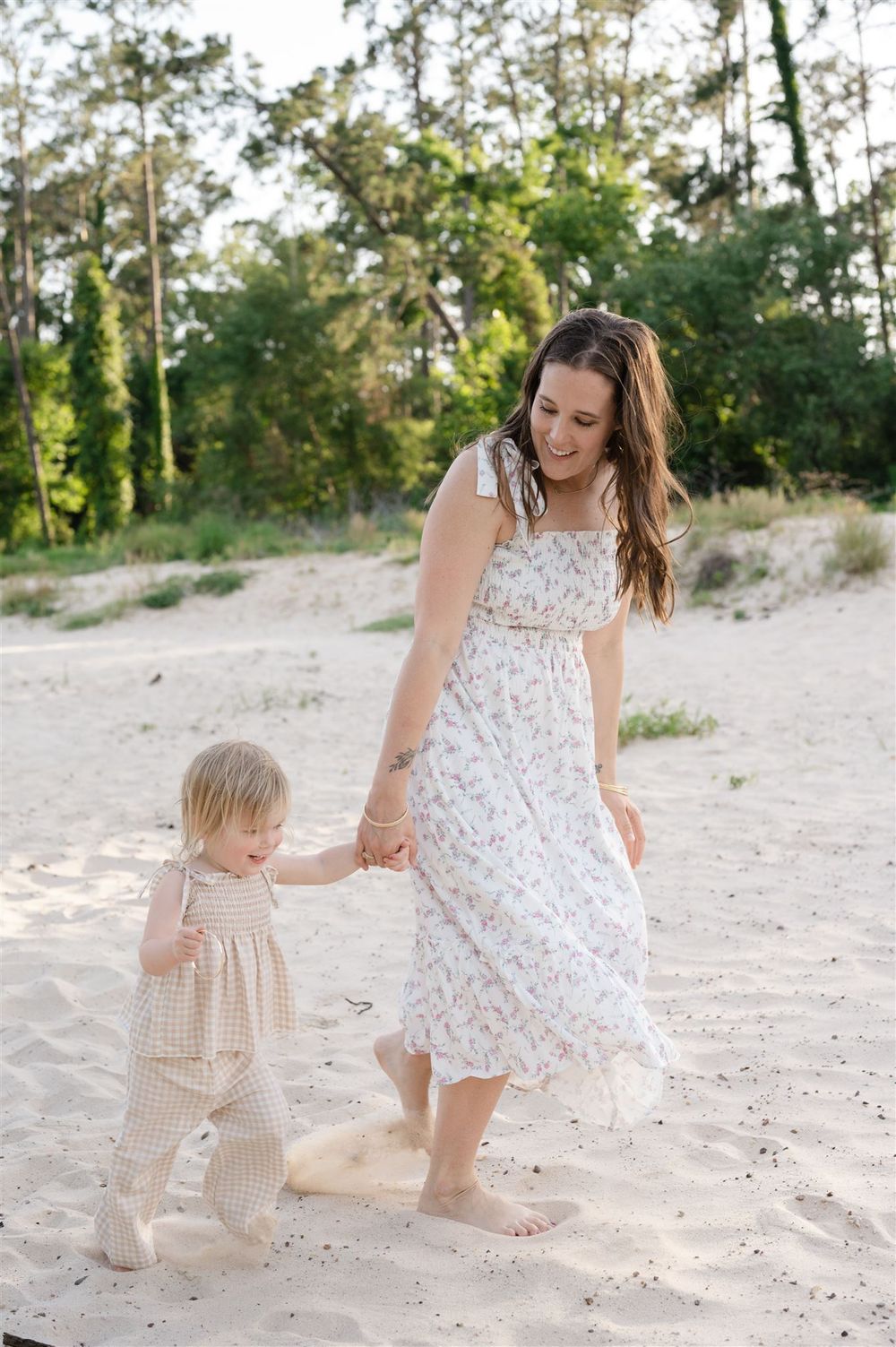 A parent and small child walk hand-in-hand on a sandy beach, both wearing light summer dresses against a wooded backdrop.