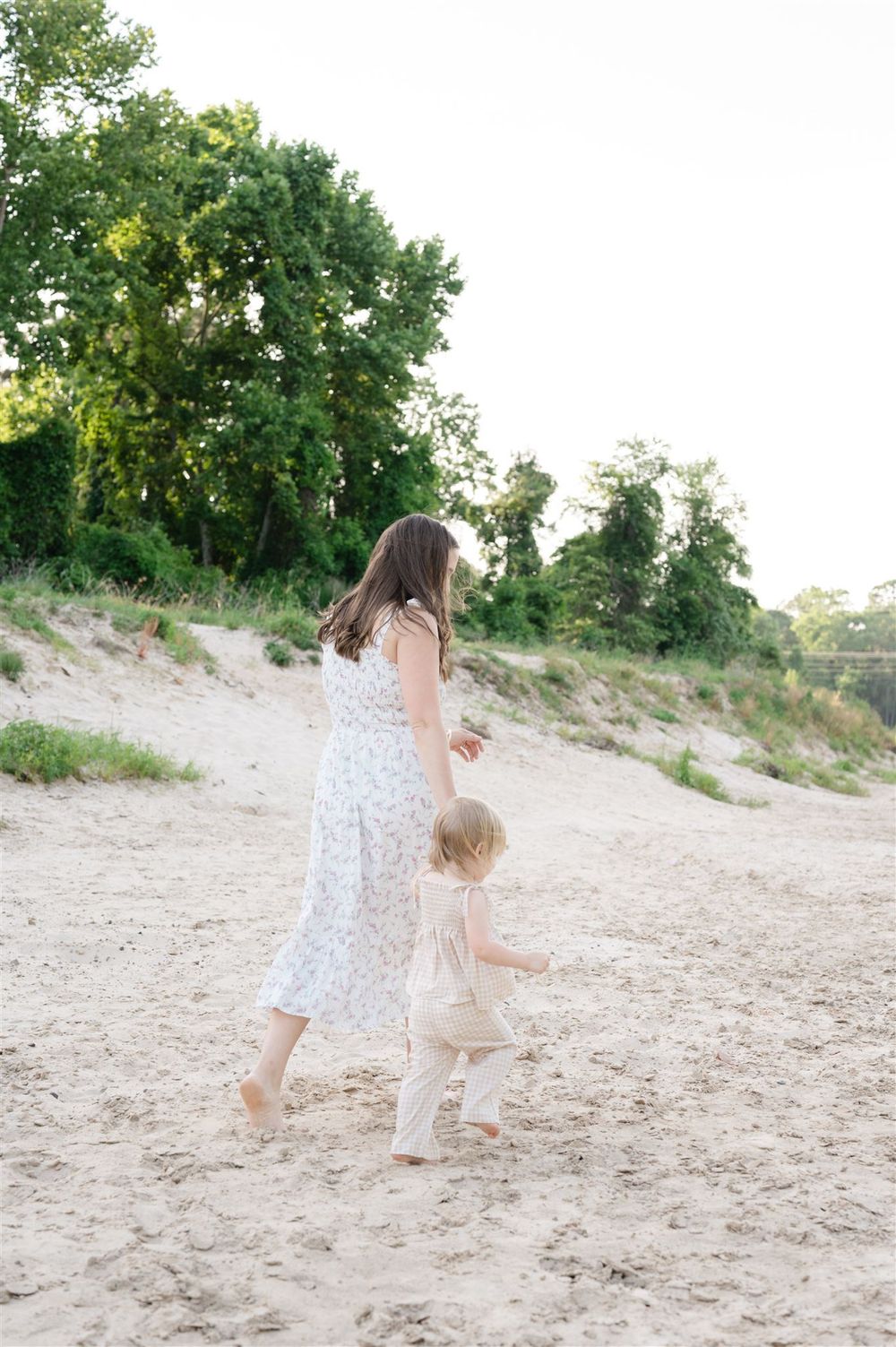 Two figures in light flowy dresses walk together on a sandy beach path bordered by green trees on a sunny day.