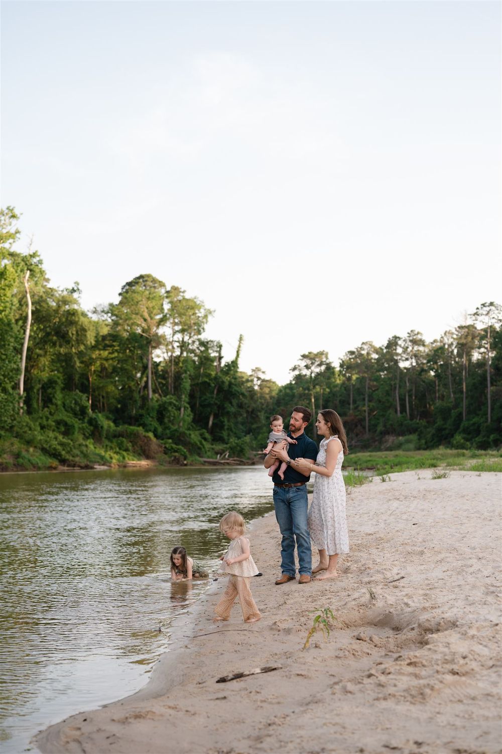 A family enjoys a peaceful afternoon on a sandy riverbank surrounded by lush forest with their dog.
