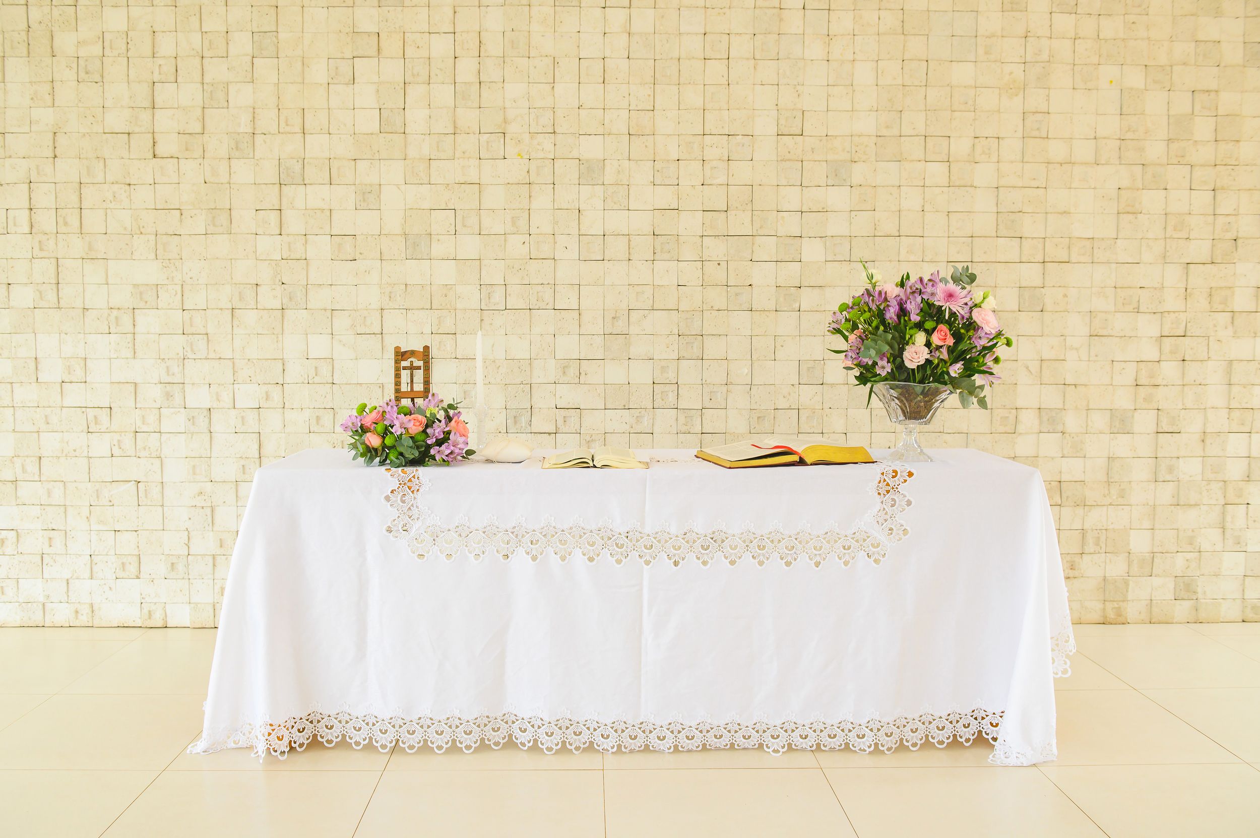 Elegant white tablecloth-covered altar with floral arrangements on a simple light-colored backdrop.