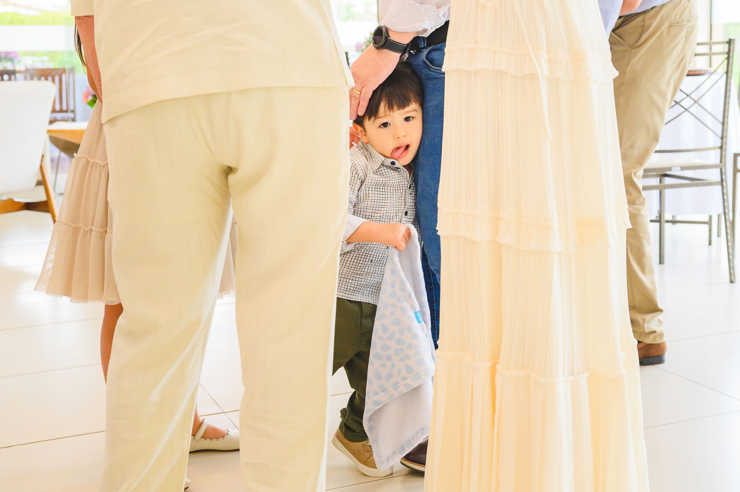 A small child peeks playfully from between adults wearing light-colored clothing in a bright indoor setting.