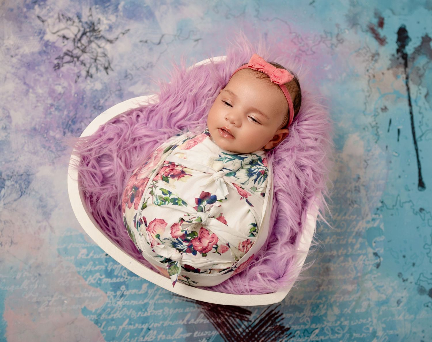 A sleeping baby lies swaddled in a floral wrap on a heart-shaped prop with purple fur against a blue background.