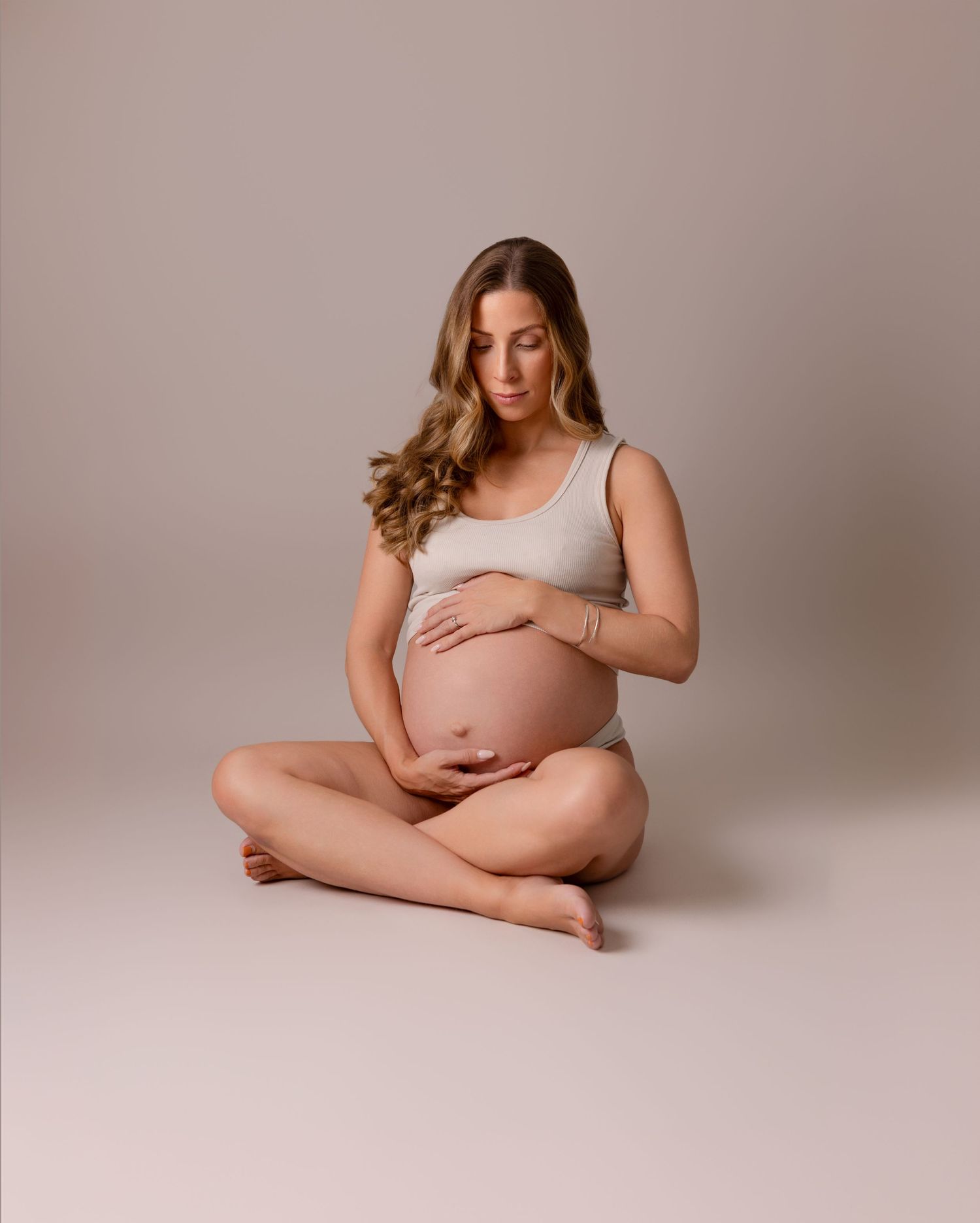 Peaceful maternity portrait of woman in beige tank top sitting cross-legged on floor.