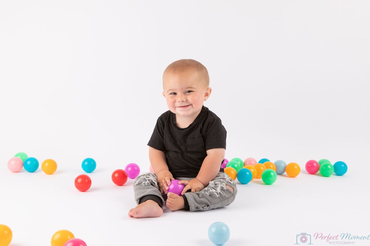 Baby in black t-shirt and camo pants sits on white floor surrounded by colorful plastic balls.