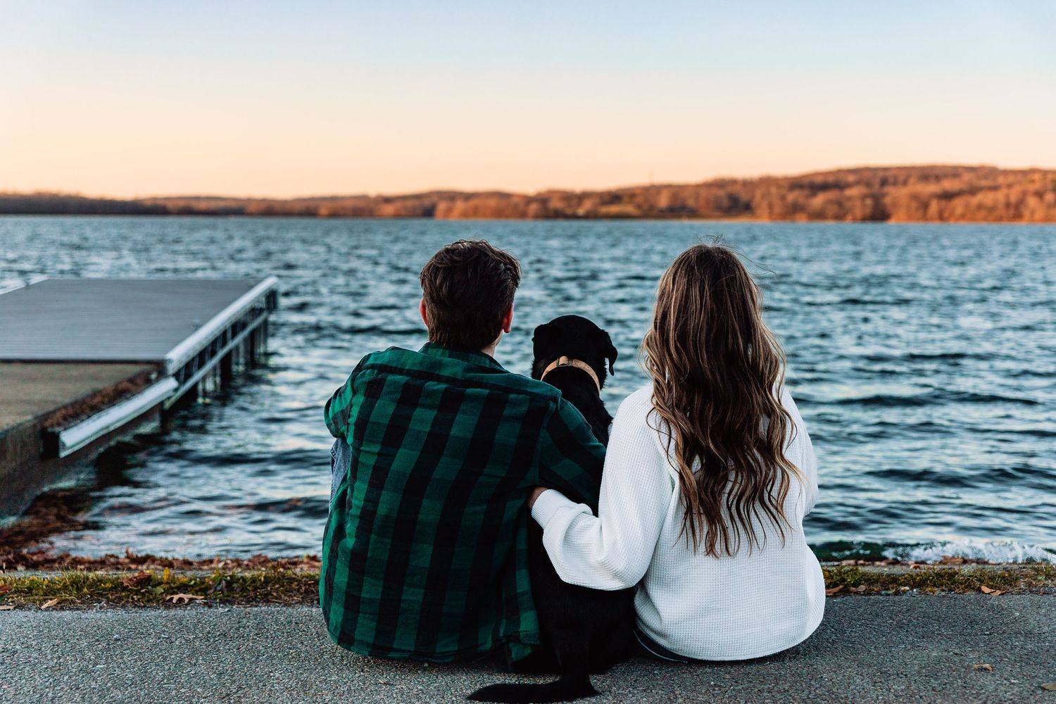 Two people sit with a dog on a dock overlooking a serene lake at sunset with hills visible in the distance.
