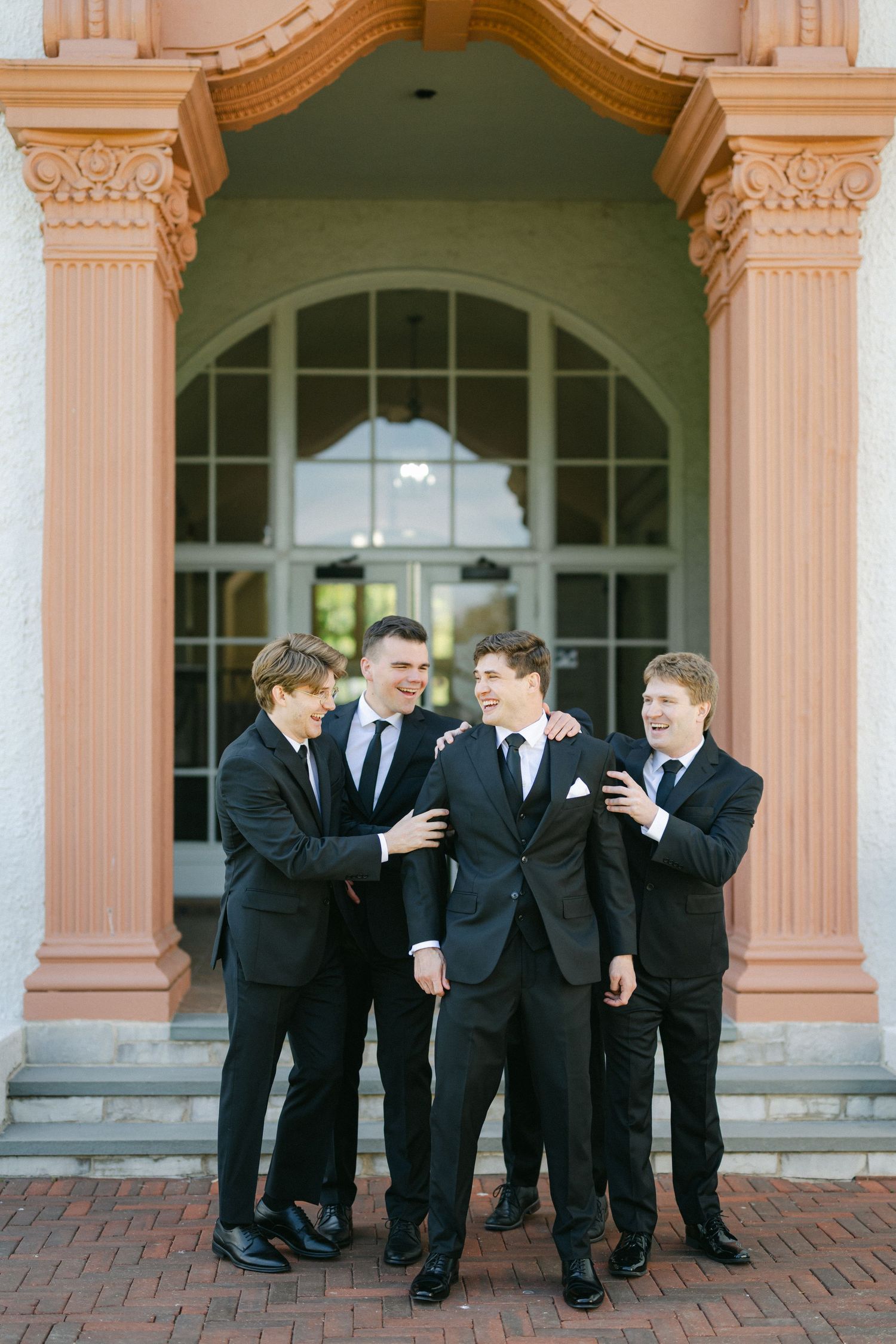 Groomsmen in black tuxedos share a candid moment and laughter on brick steps outside an elegant building.