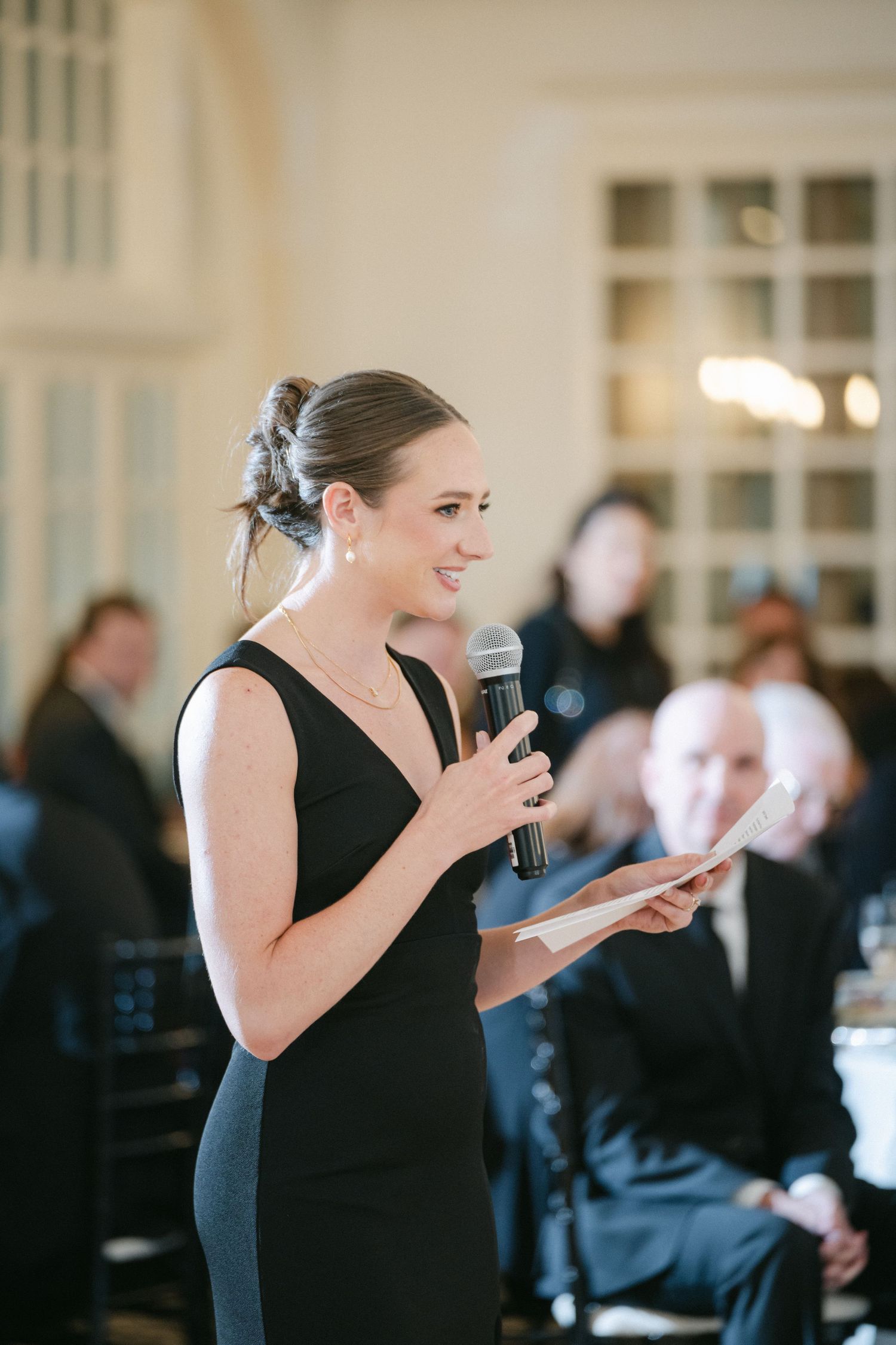 Person in elegant black dress gives speech or makes toast at formal event while holding microphone and notes.