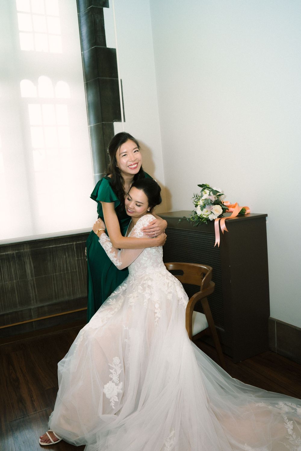 Two women embrace emotionally in wedding attire against white wall with bouquet.
