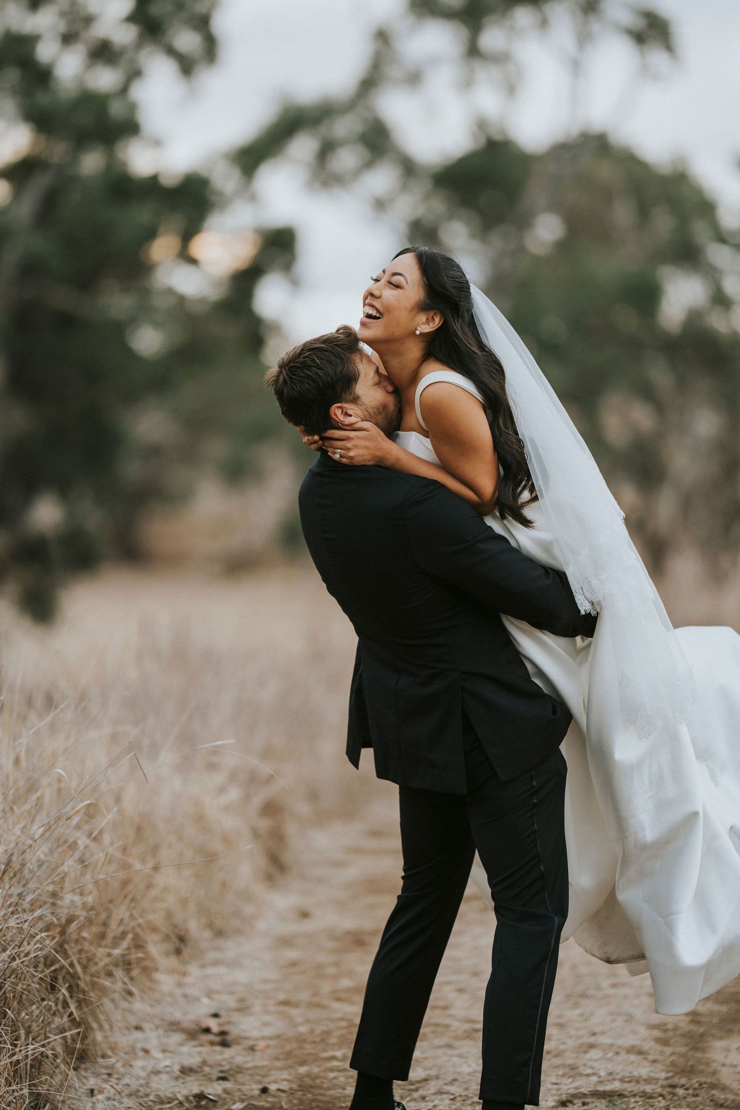 A joyful couple embraces on a dirt path during their outdoor wedding photoshoot in a dreamy natural setting.