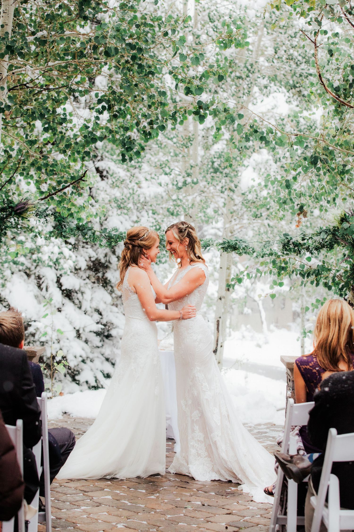 Two brides share an intimate moment at an outdoor garden ceremony surrounded by greenery and white floral decor. Alta Club. Alta, UT.