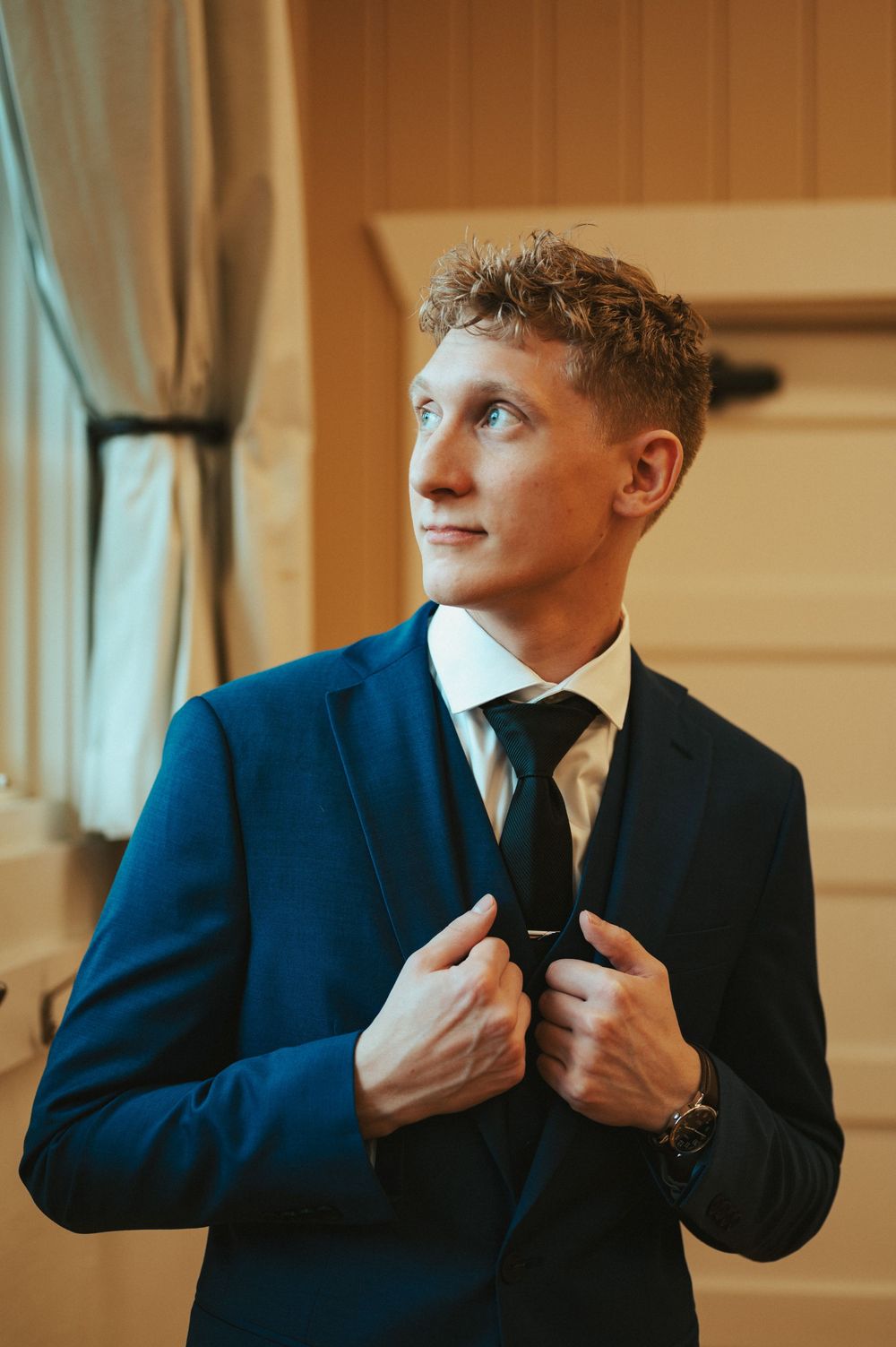 A person in a navy blue suit and tie adjusts their jacket while looking to the side in warm indoor lighting.
