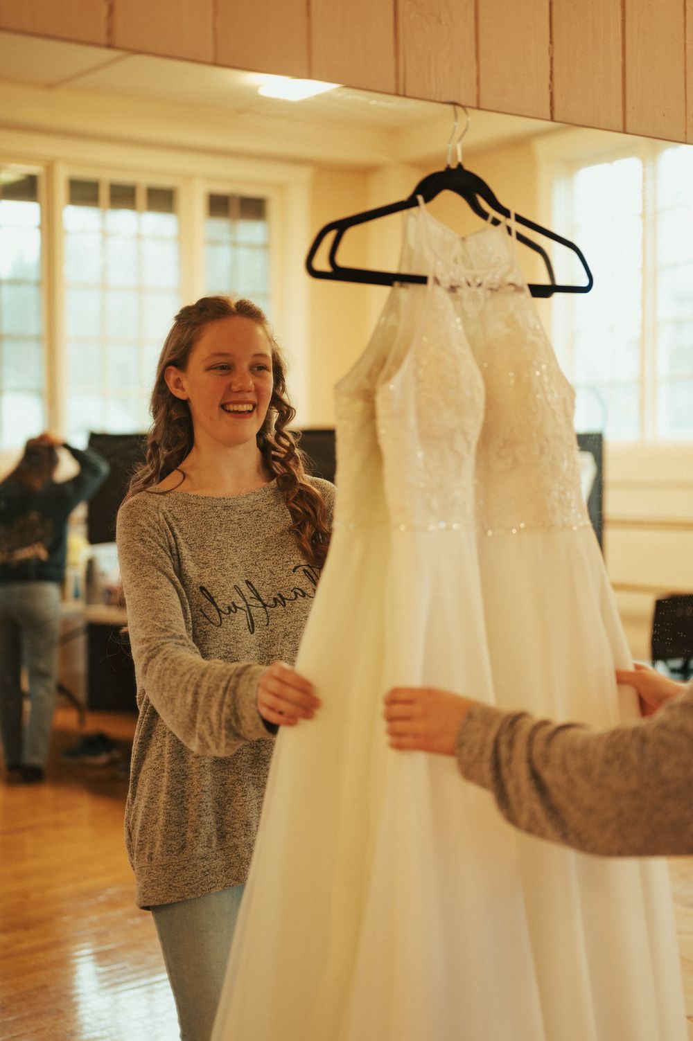 A beautiful white lace wedding dress hangs on display as someone in a gray sweater admires it during a bridal fitting.