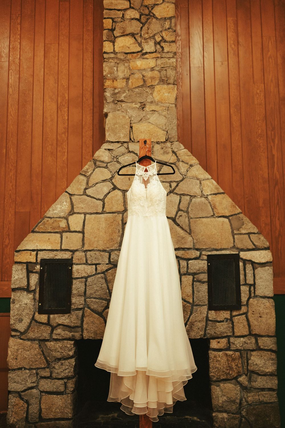 A white wedding dress hangs on a stone fireplace wall with wooden paneling on either side.