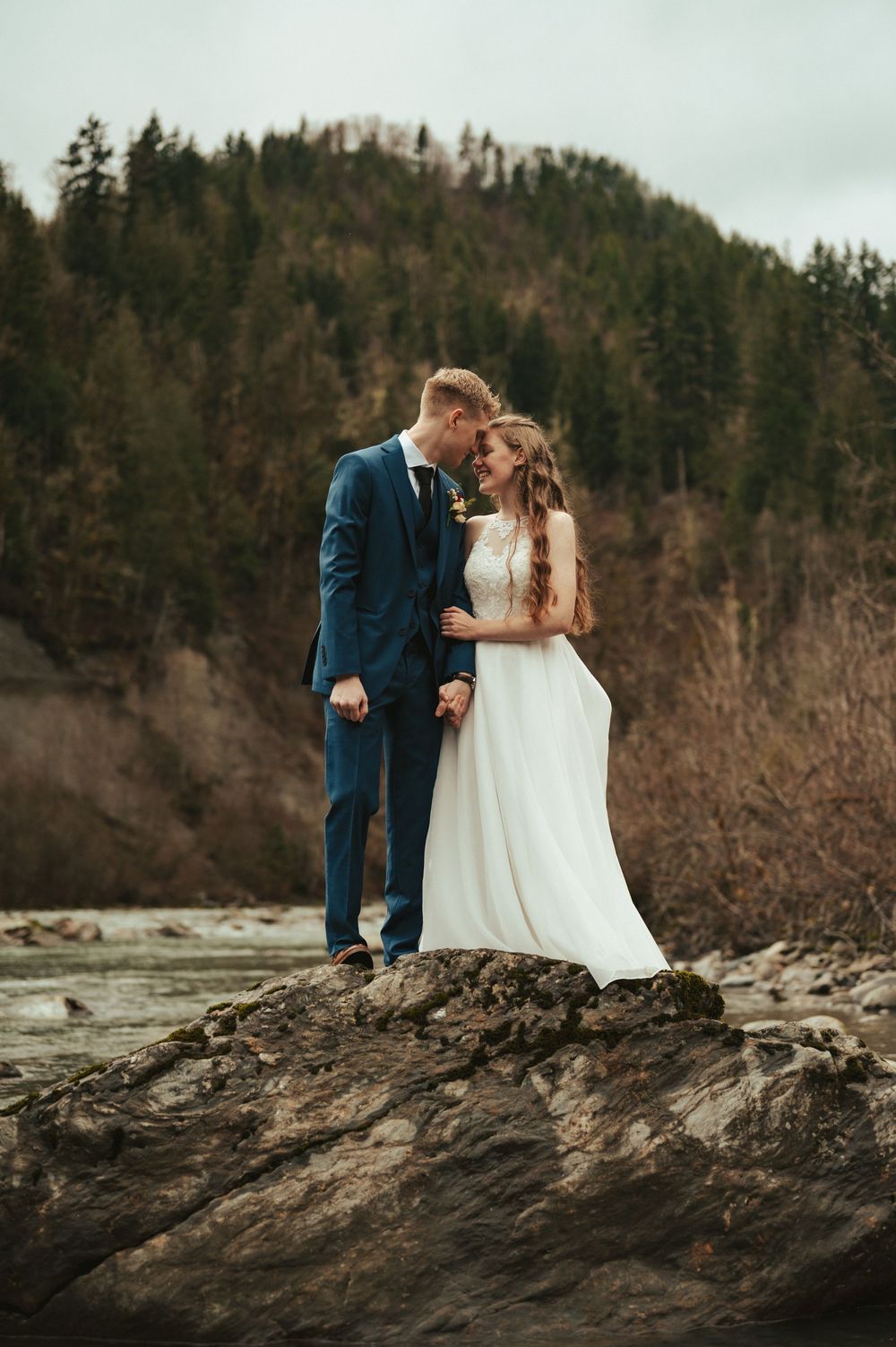 A couple in wedding attire embrace on a rocky outcrop with evergreen forested hills in the background.